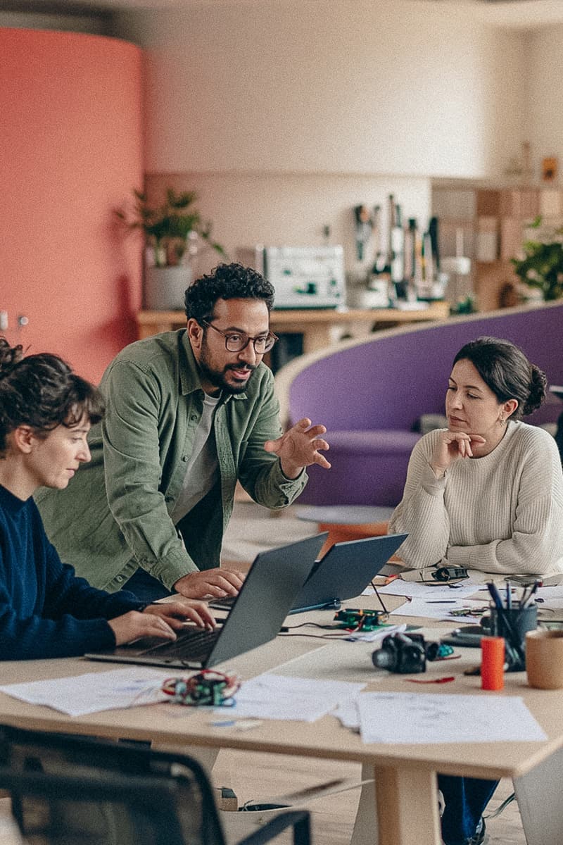 Three people collaborating around a table with laptops and prototypes